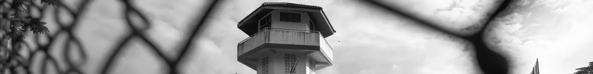 ALT text: Black and white photo of a watchtower behind a chain-link fence, highlighting security and surveillance features at a correctional facility.