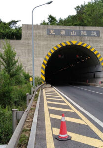 Entrance to the Harbin Expressway tunnel with safety cones and road markings, surrounded by greenery and a streetlight, under a sign with Chinese characters.