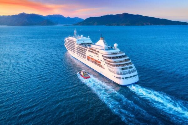 Aerial view of a large luxury cruise ship sailing in the clear blue waters of Italy, with scenic mountains in the background during sunset, highlighting a premium travel experience.