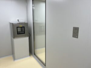 Image shows a secure access control kiosk with a touchscreen interface, located in a clean, modern office environment next to a glass door, used for visitor or employee entry management.