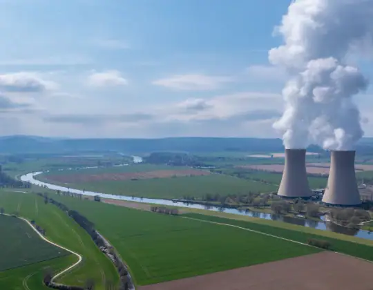 Aerial view of nuclear power plant cooling towers emitting steam over lush green fields and a river, showcasing clean energy infrastructure.