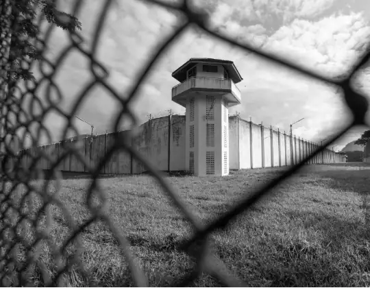 Prison watchtower viewed through a chain-link fence, symbolizing security and surveillance. The black-and-white image highlights the stark, imposing structure and the surrounding barbed wire, emphasiz.
