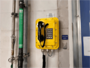 Yellow explosion-proof emergency telephone mounted on a wall in a nuclear power facility, with industrial pipes nearby.