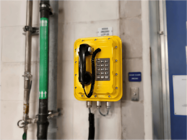 Yellow explosion-proof emergency telephone mounted on a wall in a nuclear power facility, with industrial pipes nearby.