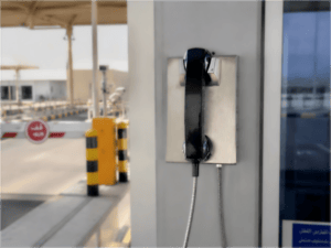 Close-up of a black public safety telephone mounted on a wall at an industrial facility in Saudi Arabia, with a parking barrier and security checkpoint in the background.