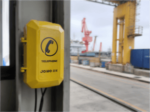 Yellow waterproof industrial telephone mounted at a shipyard with cranes and ships in the background, designed for harsh maritime environments.
