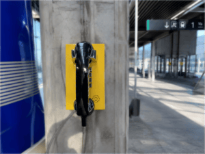 Yellow emergency telephone mounted on concrete pillar at high-speed railway station in Spain, with modern station architecture in background.