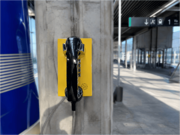 Yellow emergency telephone mounted on concrete pillar at high-speed railway station in Spain, with modern station architecture in background.